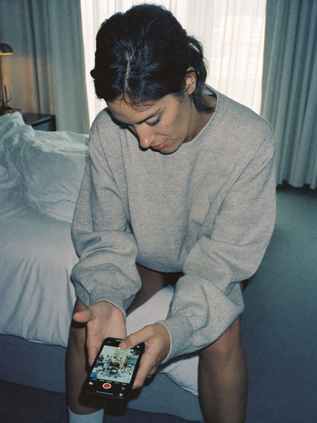 A woman alone on a hotel bed in the afternoon, looking at photos of her friends out without her.